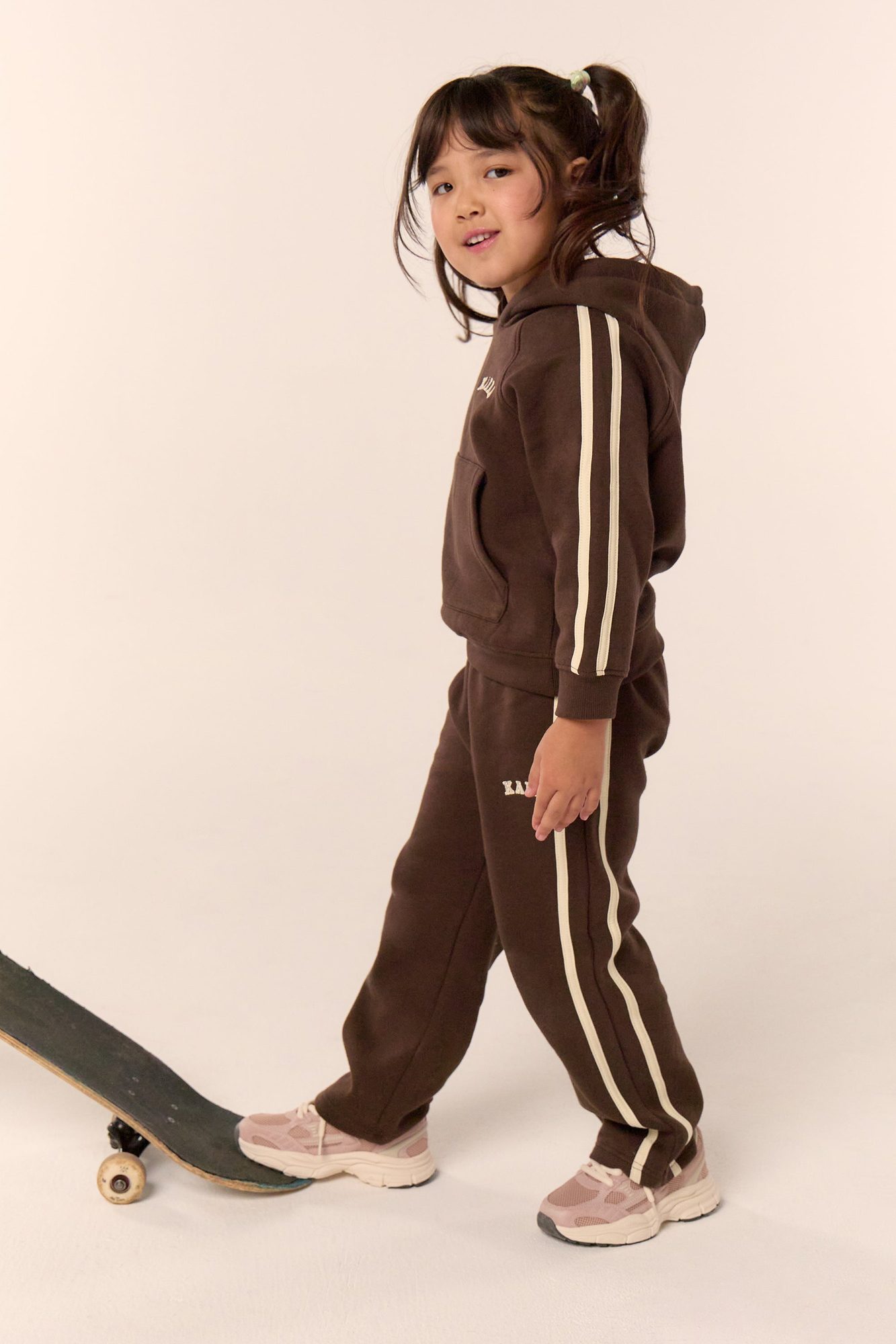 Child wearing brown hoodie and matching joggers with stripe detail, standing on skateboard