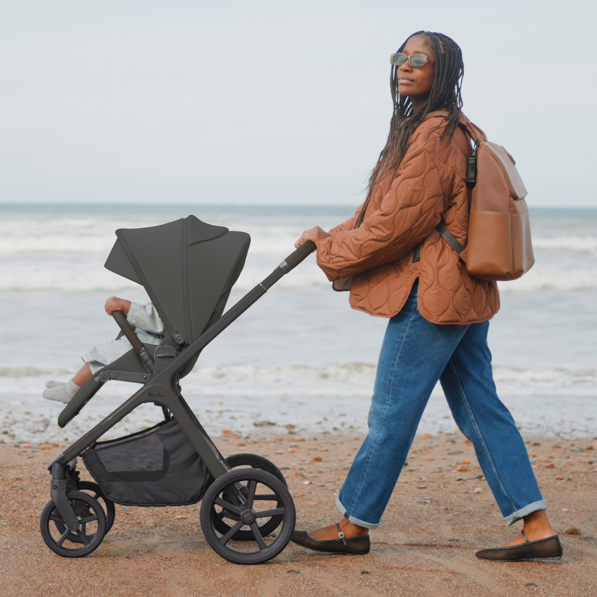 Woman with brown backpack pushing green pushchair on beach with ocean waves in background