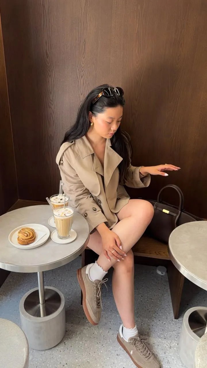 Vrouw in beige blazer zit aan tafel met koffie en gebak, dragende bruine schoenen